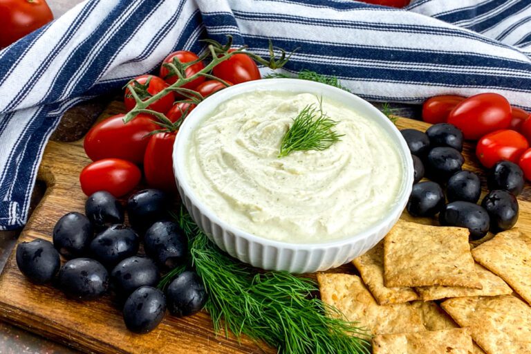 Bowl of whipped feta dip garnished with fresh dill, served with black olives and cherry tomatoes on a rustic board.