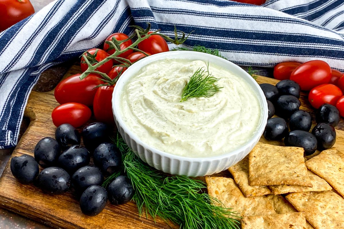 Bowl of whipped feta dip garnished with fresh dill, served with black olives and cherry tomatoes on a rustic board.