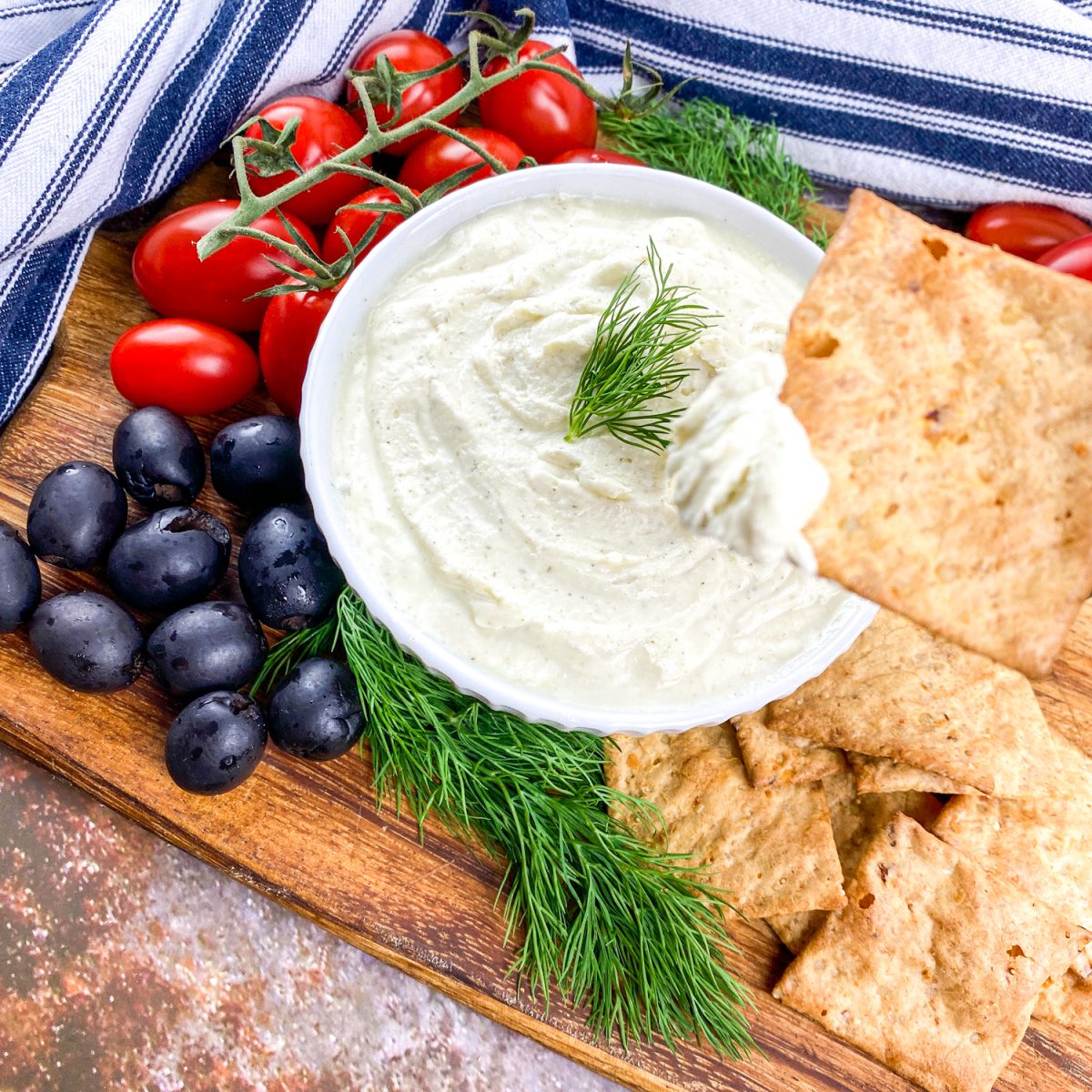 A cracker being dipped into a bowl of whipped feta dip. Garnished with fresh dill, served with black olives and cherry tomatoes on a rustic board.