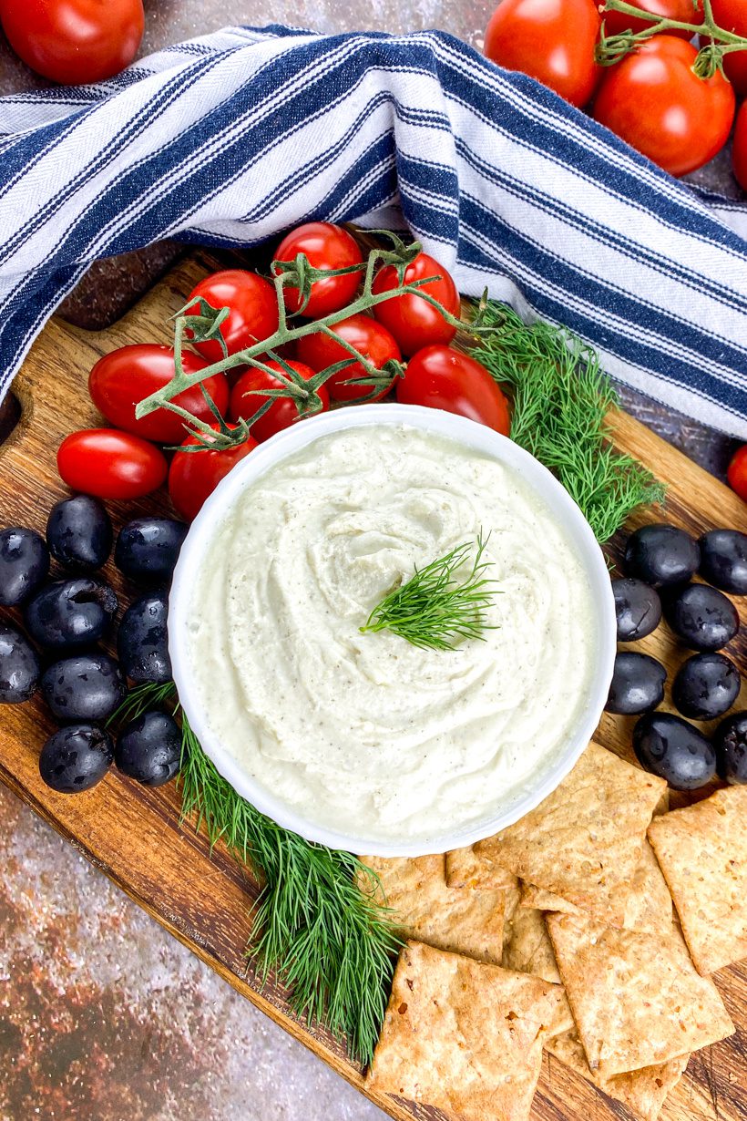 Overhead shot of bowl of whipped feta dip garnished with fresh dill, served with black olives and cherry tomatoes on a rustic board.