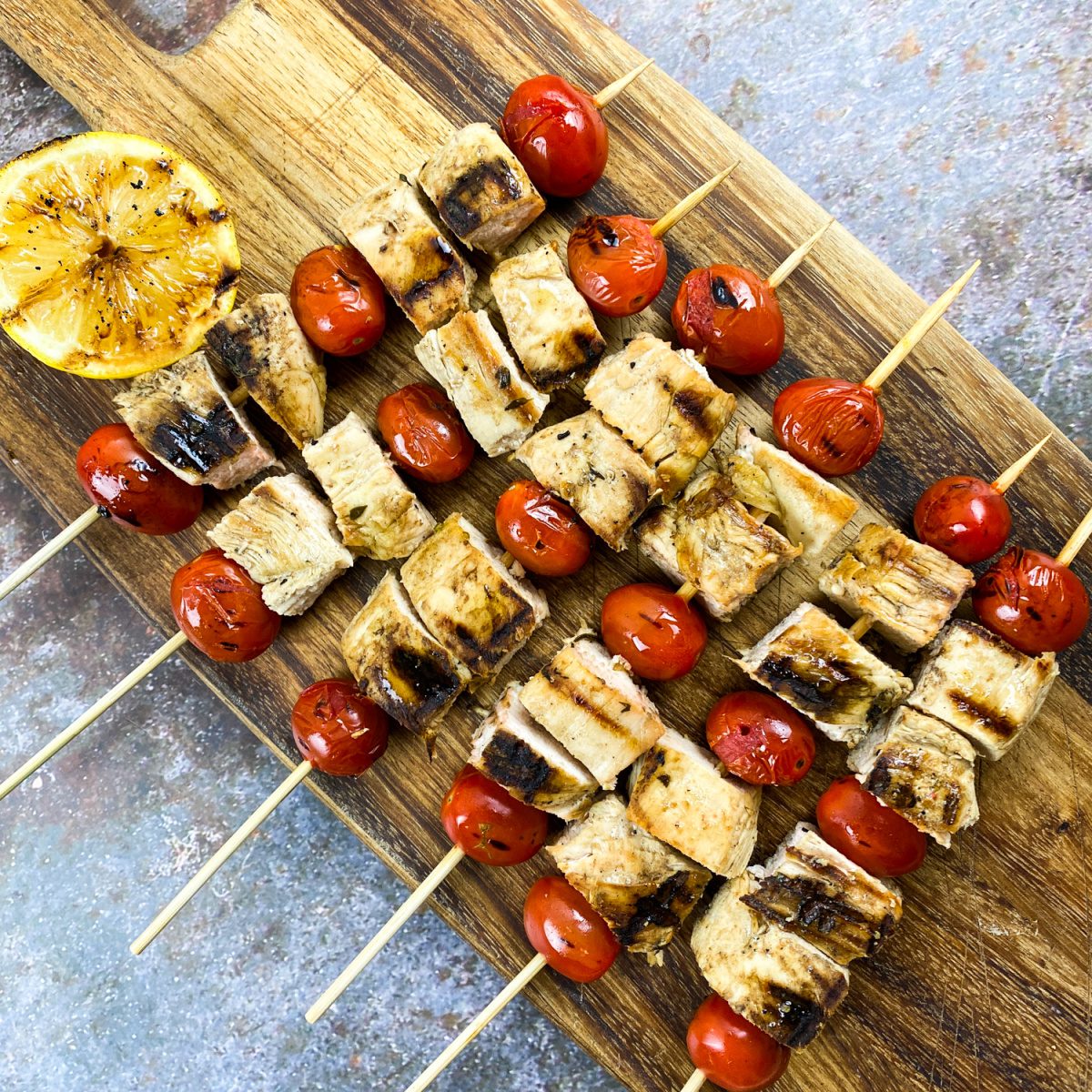 Grilled Greek chicken skewers with tomatoes and lemon on a wood platter